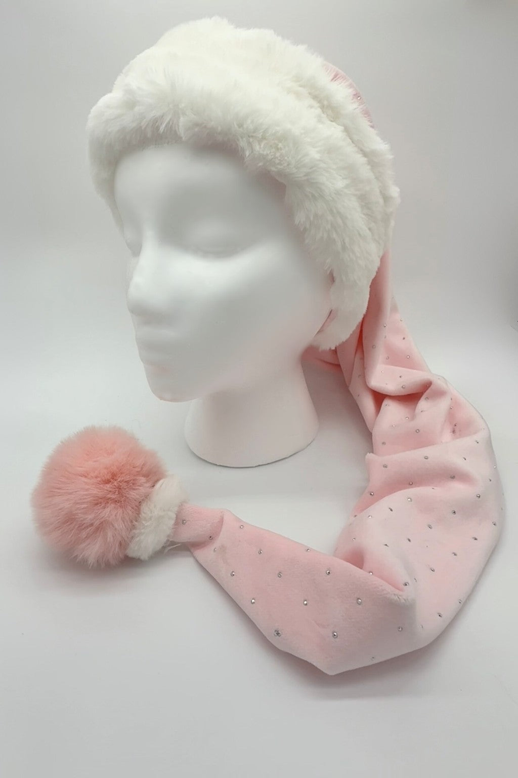 A long pink Velvet Santa-style hat with small silver rhinestones, featuring a fluffy white fur brim and a large soft pink pom-pom, displayed on a white mannequin head. There is a small white fur accent on the top of the pom-pom.