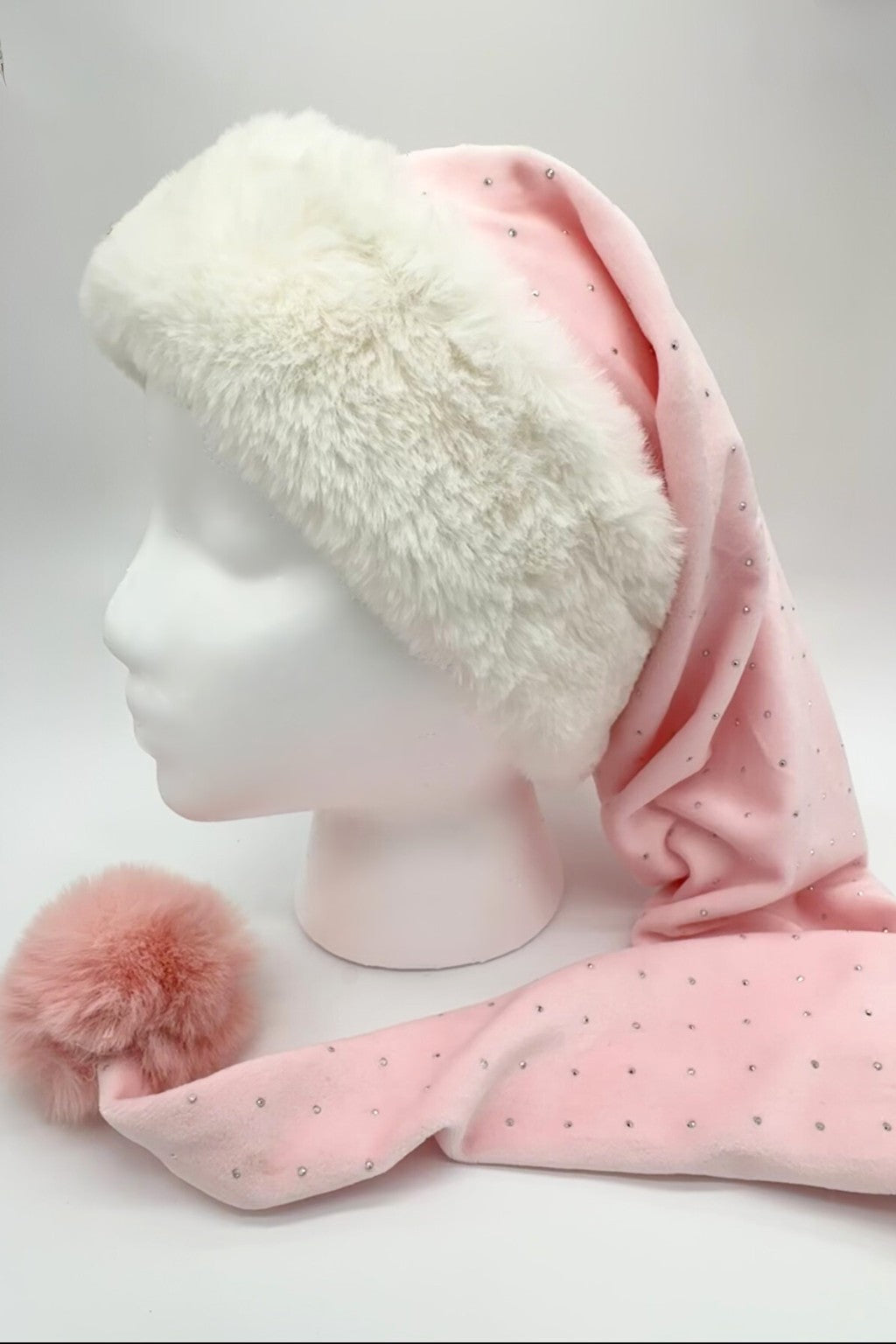 A light pink Velvet Santa-style hat decorated with small rhinestones, featuring a wide white faux fur brim and a fluffy pink pom-pom at the end. The hat is displayed on a white mannequin head against a neutral background.