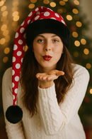 A smiling woman in a white cable-knit sweater playfully blows a kiss while wearing a long red Santa-style hat with large white polka dots, trimmed in black faux fur and ending in a black pom-pom that drapes about 8 inches below her shoulder. Warm golden holiday lights and a Christmas tree glow softly in the blurred background.