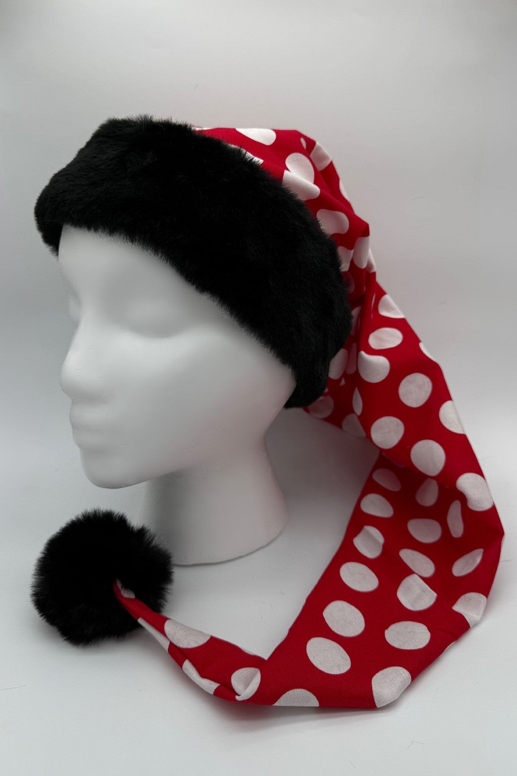 A mannequin head wearing a long red hat with large white polka dots, trimmed with black faux fur at the brim and a black pom-pom at the tip.