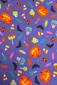 A playful Halloween-themed fabric pattern with a purple background featuring smiling jack-o’-lanterns, candy corn, lollipops, wrapped candies, cupcakes with eyepom-poms, bats, and ice cream cones, all illustrated in bright, cheerful colors with cute cartoon faces.