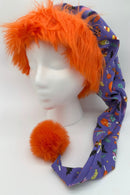 A mannequin head wearing a whimsical Halloween-themed hat. The hat has a shaggy bright orange faux fur trim and a long purple fabric body decorated with festive prints such as pumpkins, candy corn, bats, and ghosts. At the end of the hat hangs a large fluffy orange pom-pom.