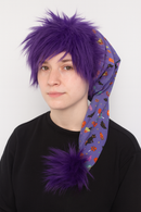A young person wearing a festive purple Halloween-themed hat with long fabric featuring pumpkins, bats, and candy prints. The hat has fluffy purple faux fur trim and two large purple faux fur poms at the end. The model is dressed in a black sweater.