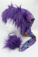 A mannequin head wearing a whimsical hat with long purple faux fur trim and a matching fluffy pom. The hat fabric is purple with colorful Halloween-themed prints, including pumpkins, bats, and candy, and it extends into a long tail ending in the large furry pom.