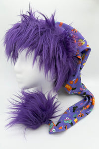 A mannequin head wearing a whimsical hat with long purple faux fur trim and a matching fluffy pom. The hat fabric is purple with colorful Halloween-themed prints, including pumpkins, bats, and candy, and it extends into a long tail ending in the large furry pom.