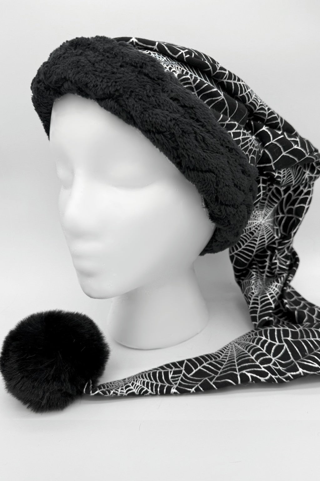 A manequin head with a black holiday hat with a fluffy black trim and a long tail featuring a white spiderweb pattern, ending in a large black pom-pom, displayed on a white mannequin head.