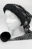 A manequin head with a black holiday hat with a fluffy black trim and a long tail featuring a white spiderweb pattern, ending in a large black pom-pom, displayed on a white mannequin head.