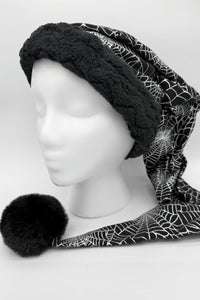 A manequin head with a black holiday hat with a fluffy black trim and a long tail featuring a white spiderweb pattern, ending in a large black pom-pom, displayed on a white mannequin head.