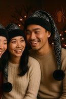 Smiling couple sitting close together, each wearing a festive black Santa-style hat with white spiderweb patterns, black faux-fur trim, and black pom-poms, in a cozy indoor holiday setting with warm lighting and seasonal decorations.