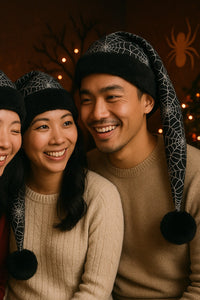 Smiling couple sitting close together, each wearing a festive black Santa-style hat with white spiderweb patterns, black faux-fur trim, and black pom-poms, in a cozy indoor holiday setting with warm lighting and seasonal decorations.