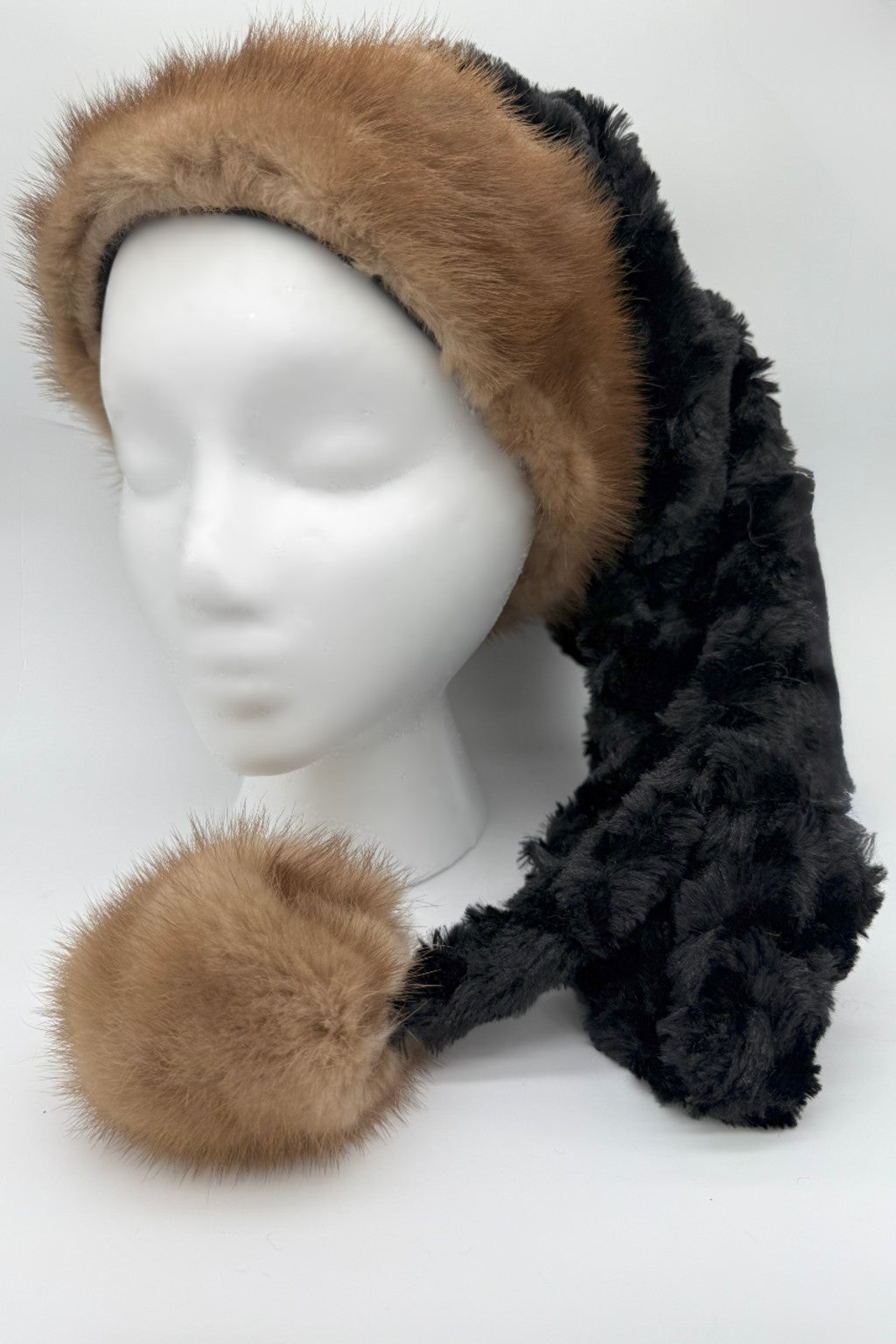 A mannequin head wearing a black panne Velvet hat with a long tail that ends in a large, fluffy pom-pom. The hat has a soft, textured pattern and a slightly shaggy look, styled in a cozy, winter fashion. The fur on the trim and pom-pom is real fur.