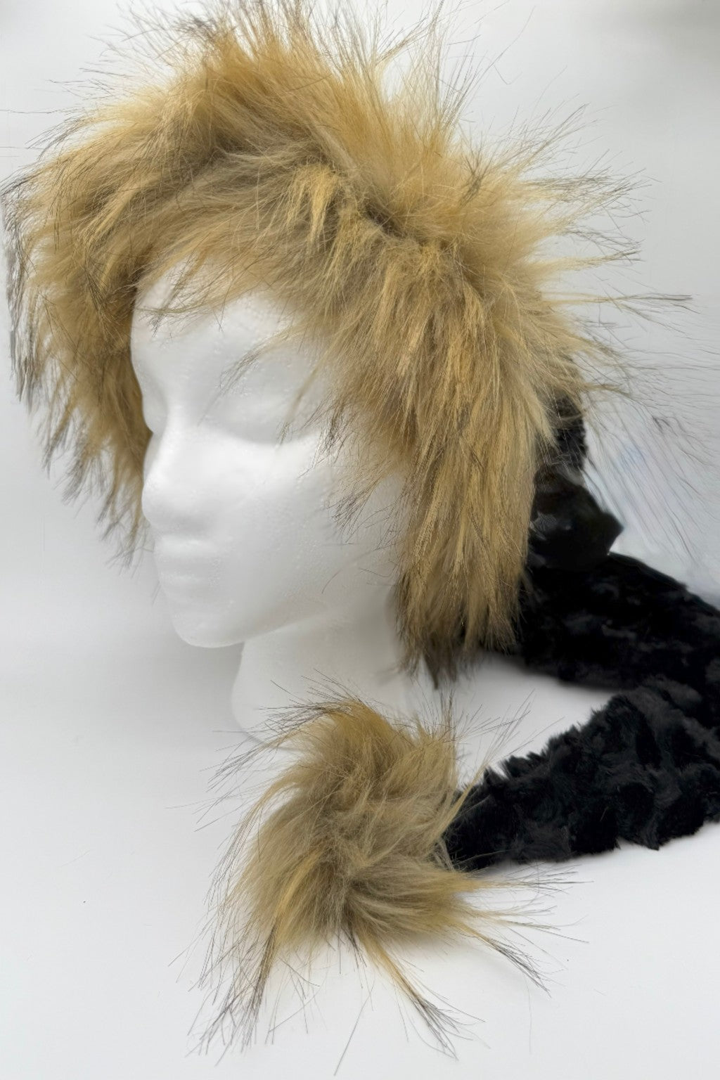 A mannequin head displays a black panne Velvet hat with long tan faux fur trim and a matching tan faux fur pom-pom at the end of its tail-like extension.
