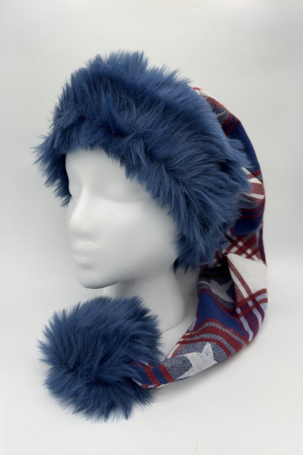 A mannequin head wearing a long festive hat with a wide, fluffy blue faux fur trim and a large matching pom-pom at the end. The main body of the hat features a red, white, and blue plaid and star pattern.