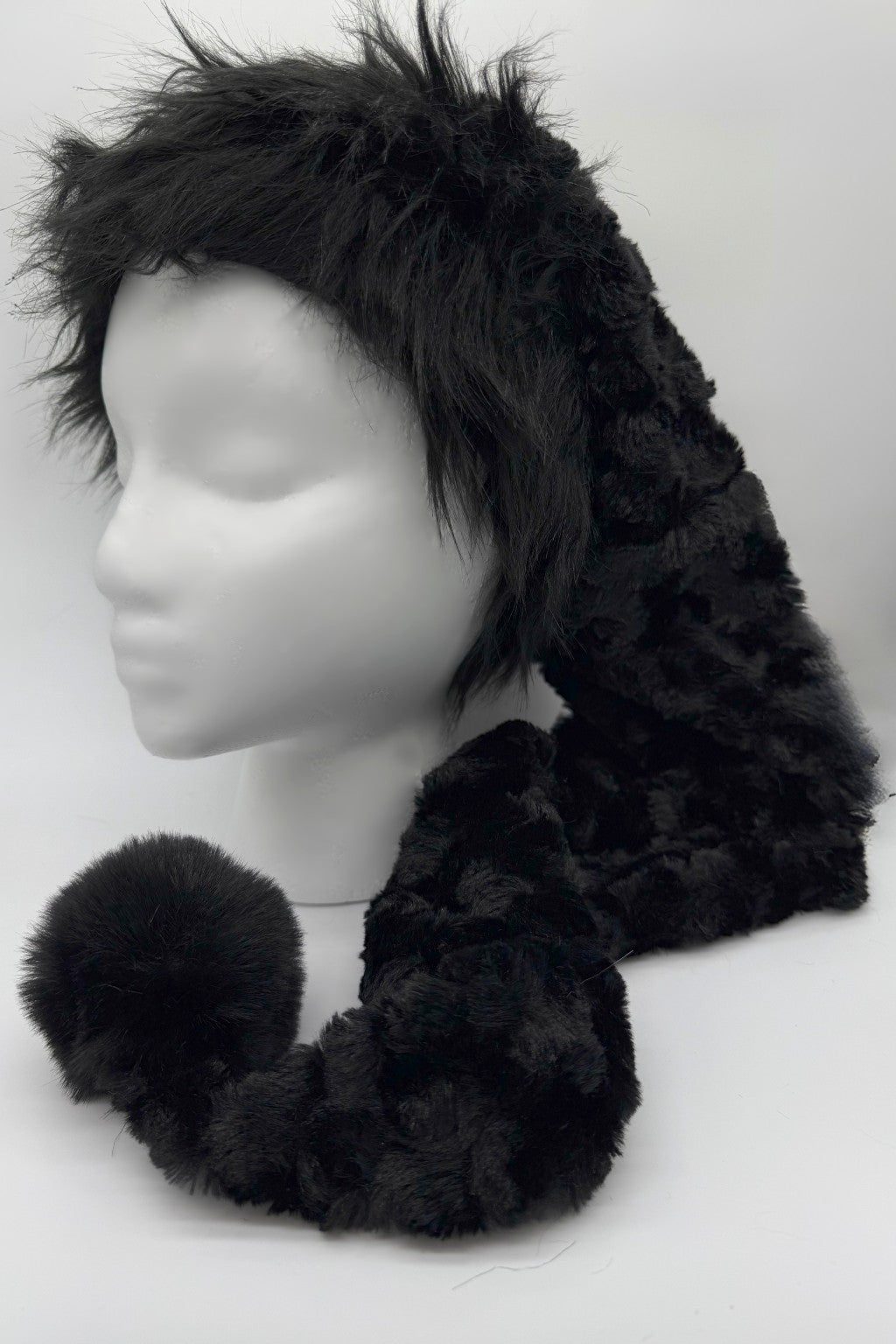 A black panne Velvet  hat with a long tail ending in a large fluffy black trim and pom-pom, displayed on a white mannequin head against a plain white background.