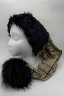 A mannequin head displays a winter hat with black faux fur trim and a beige plaid fabric (burbery) body. The hat has long scarf-like extensions ending in large black faux fur pom-poms.
