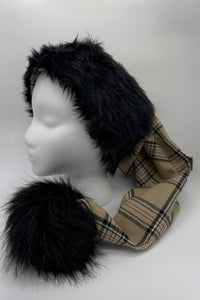 A mannequin head displays a winter hat with black faux fur trim and a beige plaid fabric (burbery) body. The hat has long scarf-like extensions ending in large black faux fur pom-poms.