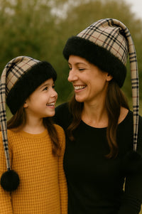 Mother and daughter wearing matching Burberry plaid hats with black faux-fur trim and black pom, smiling outdoors together.