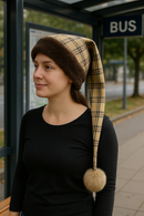 A woman wearing a long, plaid winter hat with a fur trim and pom-pom stands at a bus stop. She is dressed in a black long-sleeve shirt and is looking to the side. The bus stop sign is visible in the background.