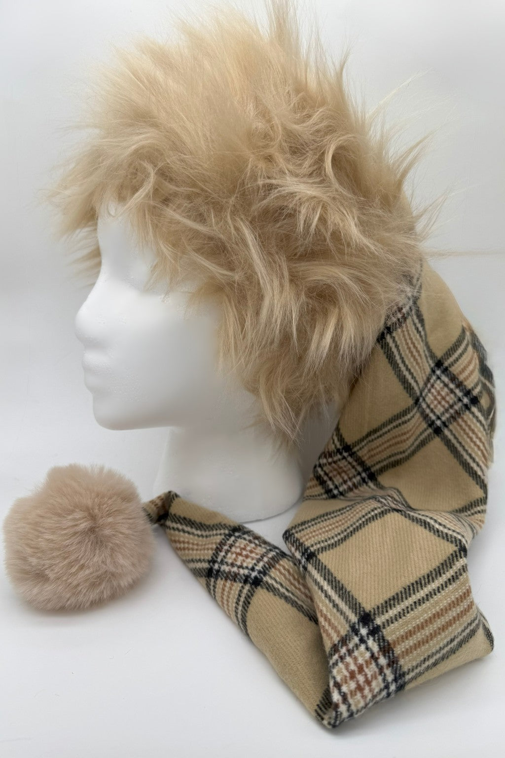 A mannequin head with shaggy, tan faux fur trimmed Santa-style hat of Burbrrry plaid fabric. A matching fluffy beige pom-pom accessory rests beside the scarf.