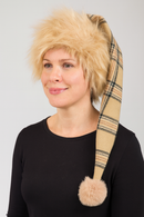  person with short blonde hair is wearing a long, plaid, beige and brown hat with a fluffy pom-pom at the end. The person is dressed in a plain black top and is smiling slightly, facing slightly to the left. The background is plain and light-colored.