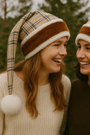 Two young women standing outdoors, smiling warmly at each other. Both are wearing matching beige plaid Santa-style hats with brown and white faux fur trim and large white pom-poms at the ends. One wears a cream sweater, and the other wears a dark brown sweater, with blurred trees in the background.