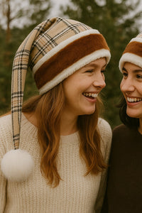Two young women standing outdoors, smiling warmly at each other. Both are wearing matching beige plaid Santa-style hats with brown and white faux fur trim and large white pom-poms at the ends. One wears a cream sweater, and the other wears a dark brown sweater, with blurred trees in the background.