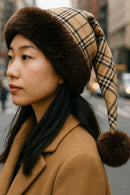 Asian model wearing a handmade Burberry-style plaid hat with brown trim and brown pom-pom, standing in a busy city street.